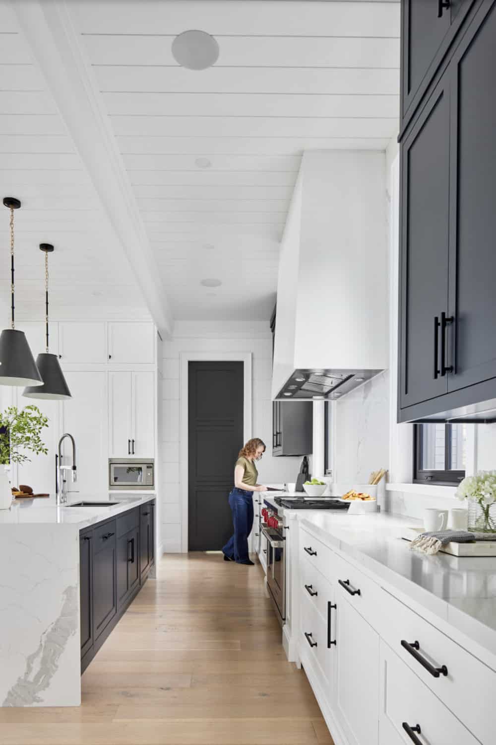 Galley kitchen view with person cooking at range, navy upper cabinets, marble backsplash, and shiplap ceiling