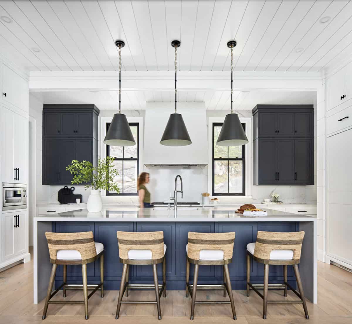 Symmetrical kitchen with navy island, four woven barstools, three black pendant lights, and marble backsplash