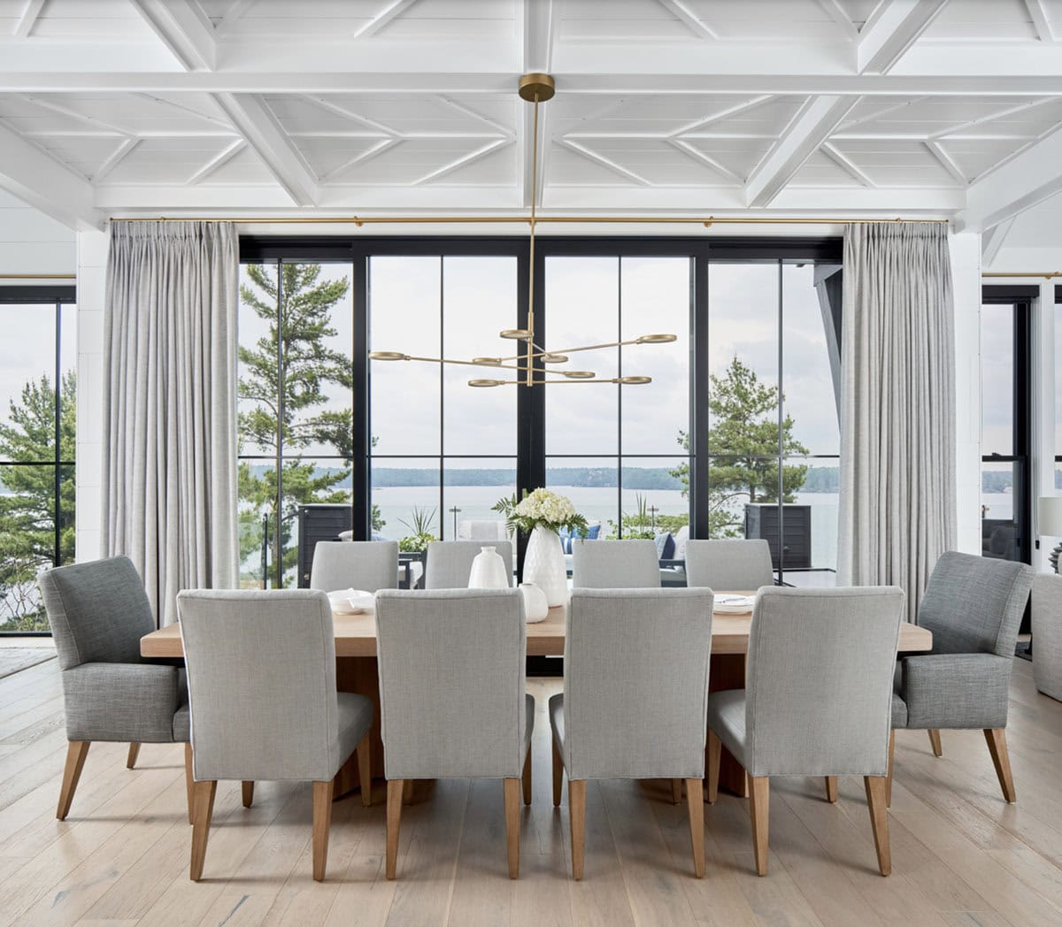 Dining room with ten grey upholstered chairs, oak table, brass linear chandelier, and panoramic lake windows