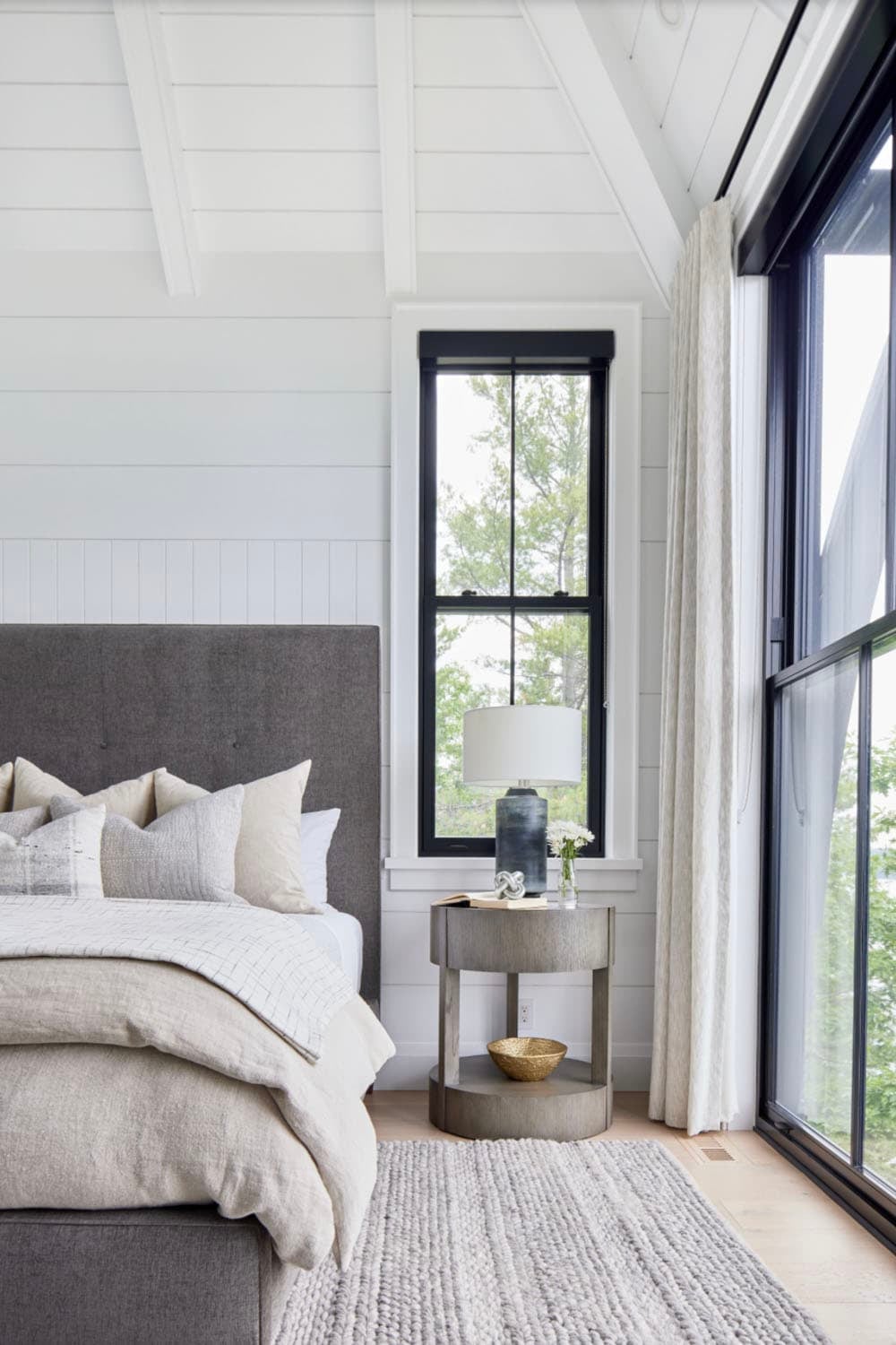 Primary bedroom corner with tufted grey headboard, round nightstand, and floor-to-ceiling black-framed windows