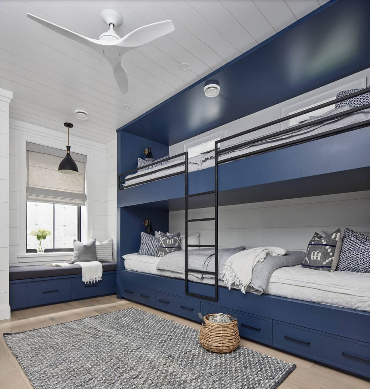 Navy built-in bunk room with four bunks, black metal ladders, window seat, and storage drawers