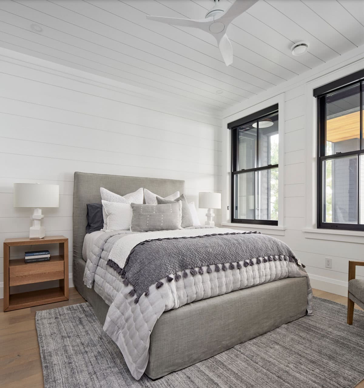 Guest bedroom with grey upholstered bed, white shiplap walls, white ceiling fan, and black-framed windows