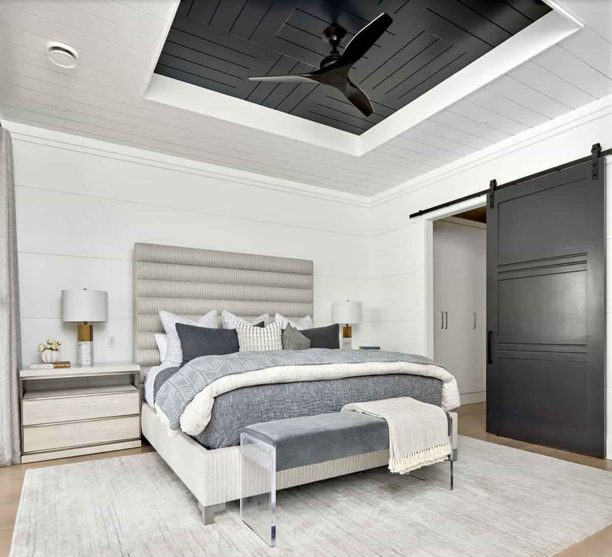 Primary bedroom with channeled headboard, black tray ceiling with fan, acrylic bench, and dark barn door