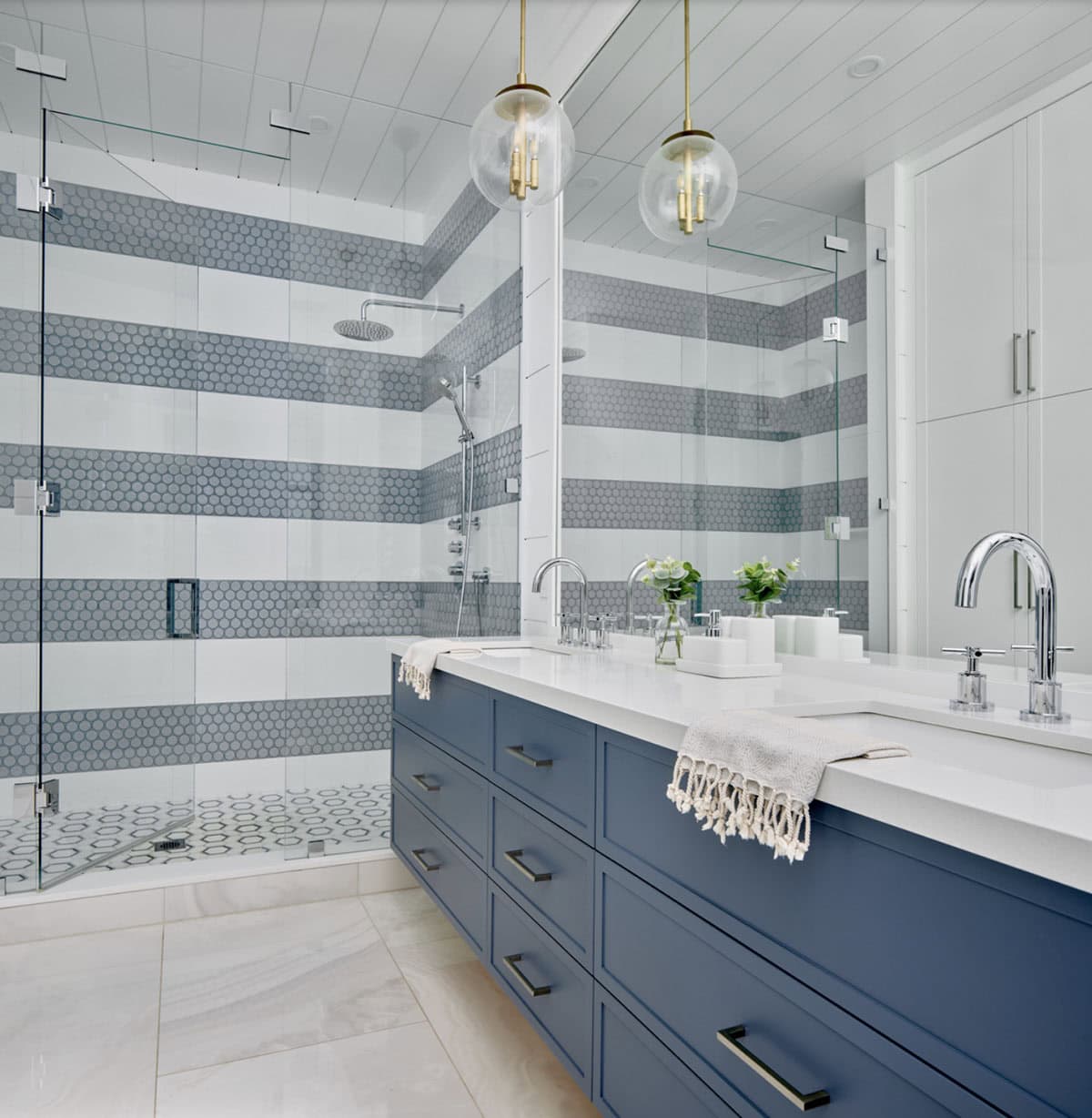 Ensuite with navy double vanity, striped penny tile shower walls, glass enclosure, and brass globe pendants