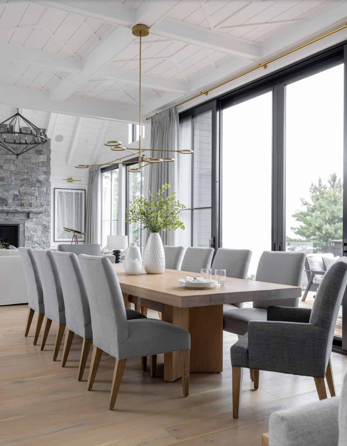 Cottage dining room with long oak table, grey upholstered chairs, brass ring chandelier, and lake-view doors