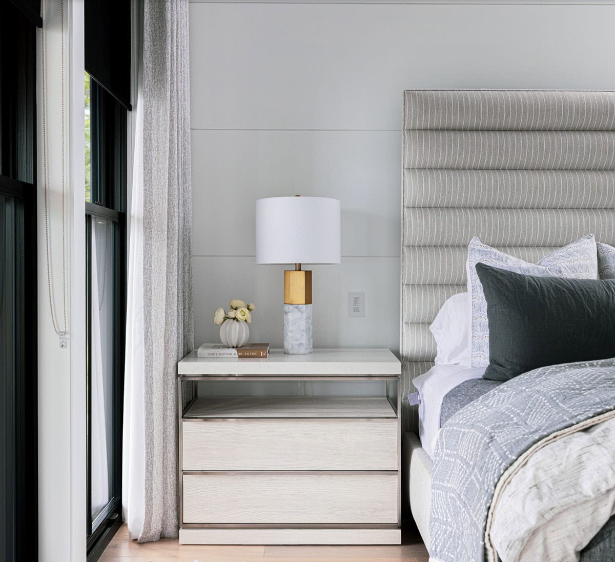 Primary bedroom nightstand detail with marble and brass lamp, white ceramic vase, and channeled headboard