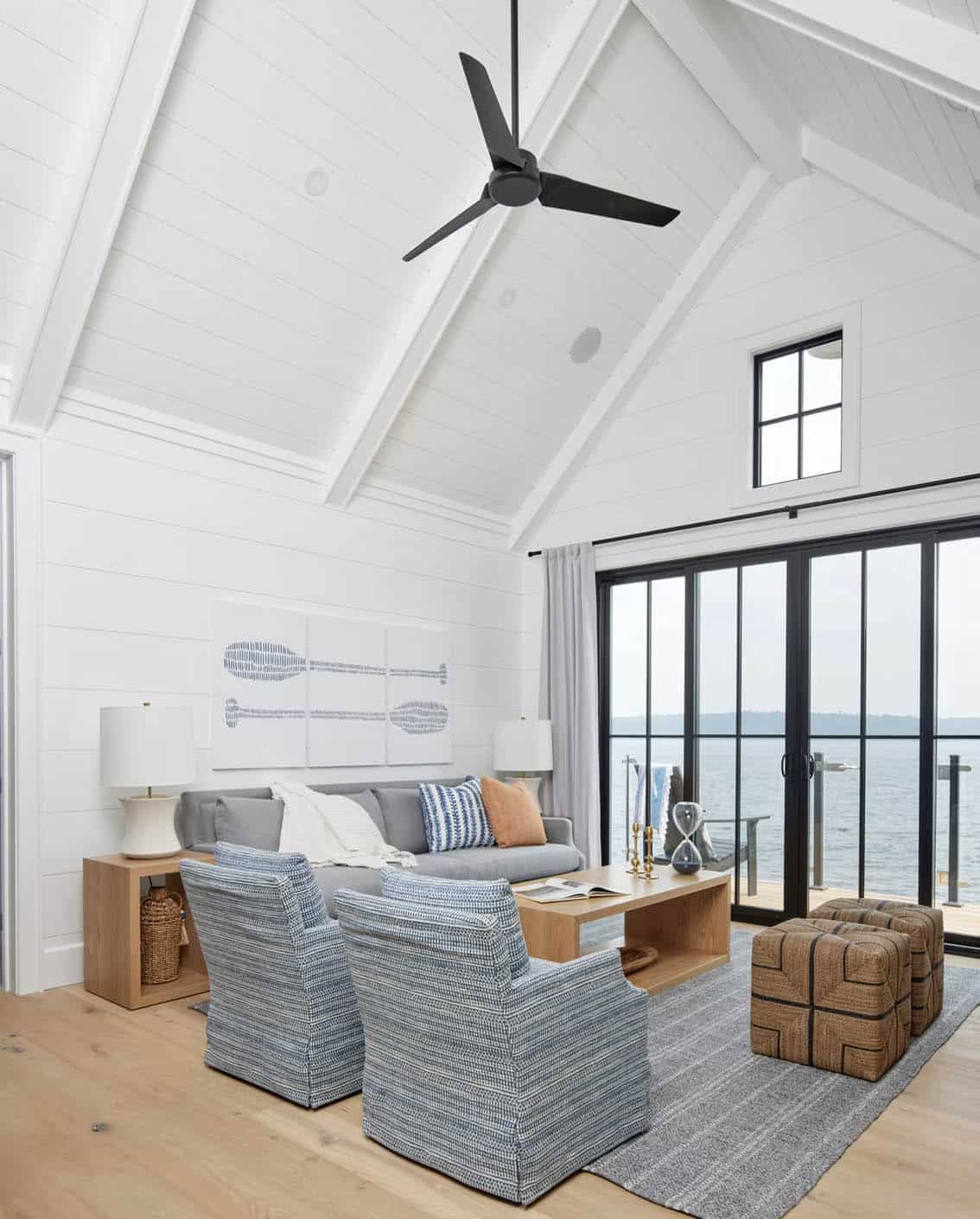 Boathouse living room with vaulted shiplap ceiling, black ceiling fan, gray sofa, and lake views through black-framed glass doors