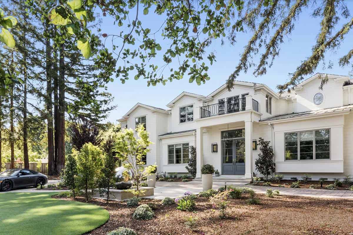White two-story transitional estate with gray front door, iron balcony railing, and mature redwood trees