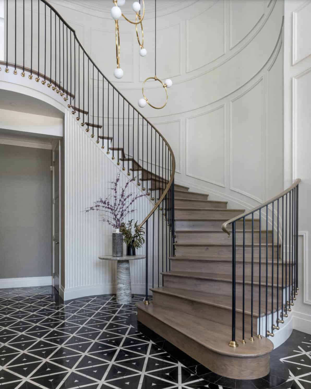 Curved staircase with black iron balusters, brass accents, and geometric black-and-white marble foyer floor