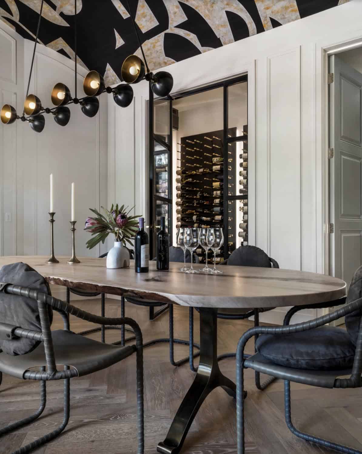 Close-up of live-edge dining table with rebar-frame chairs and backlit wine cellar behind steel-framed glass door