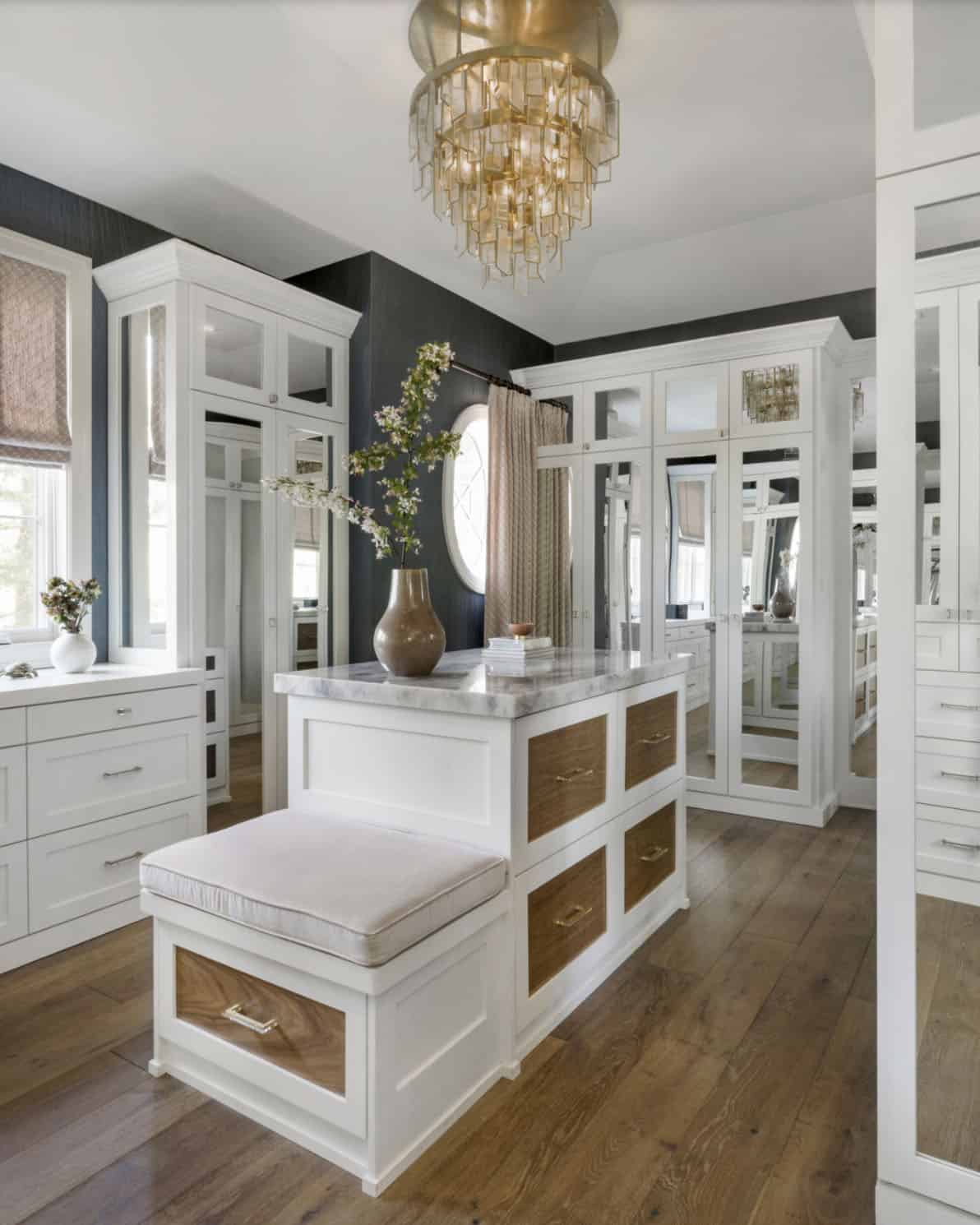 Primary walk-in closet with white cabinetry, mirrored wardrobe doors, marble island, and brass chandelier