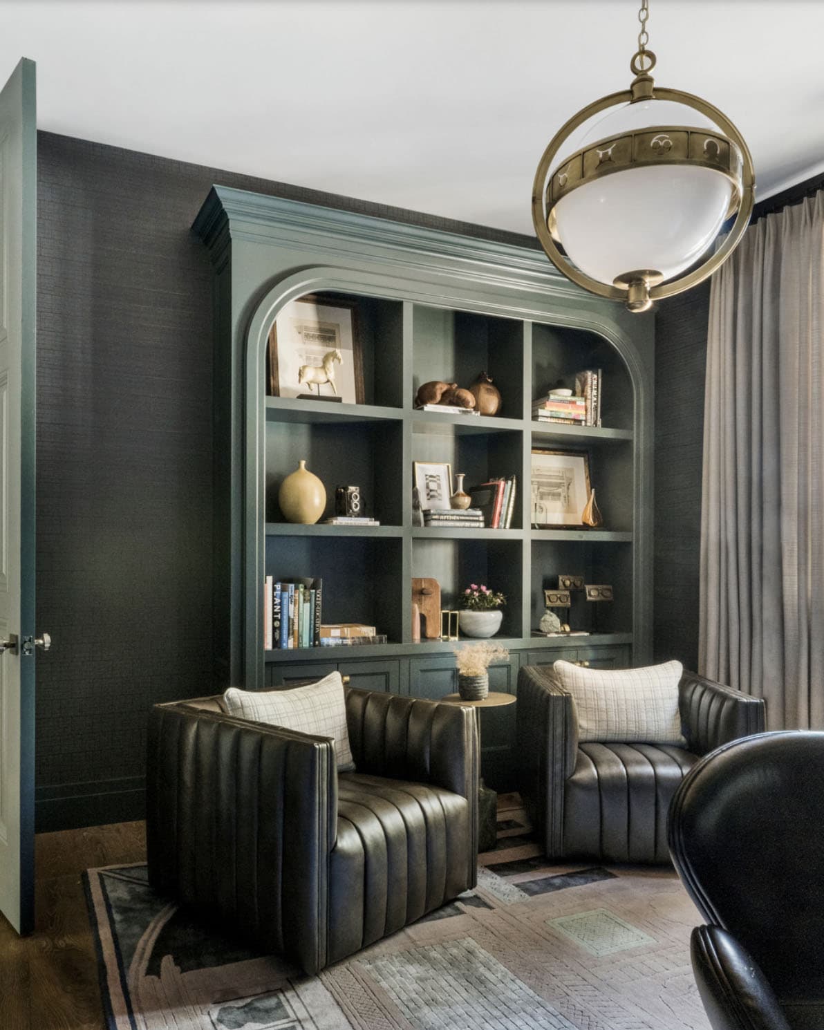 Dark home office with sage green arched built-in bookcase, channel-tufted leather chairs, and brass globe pendant