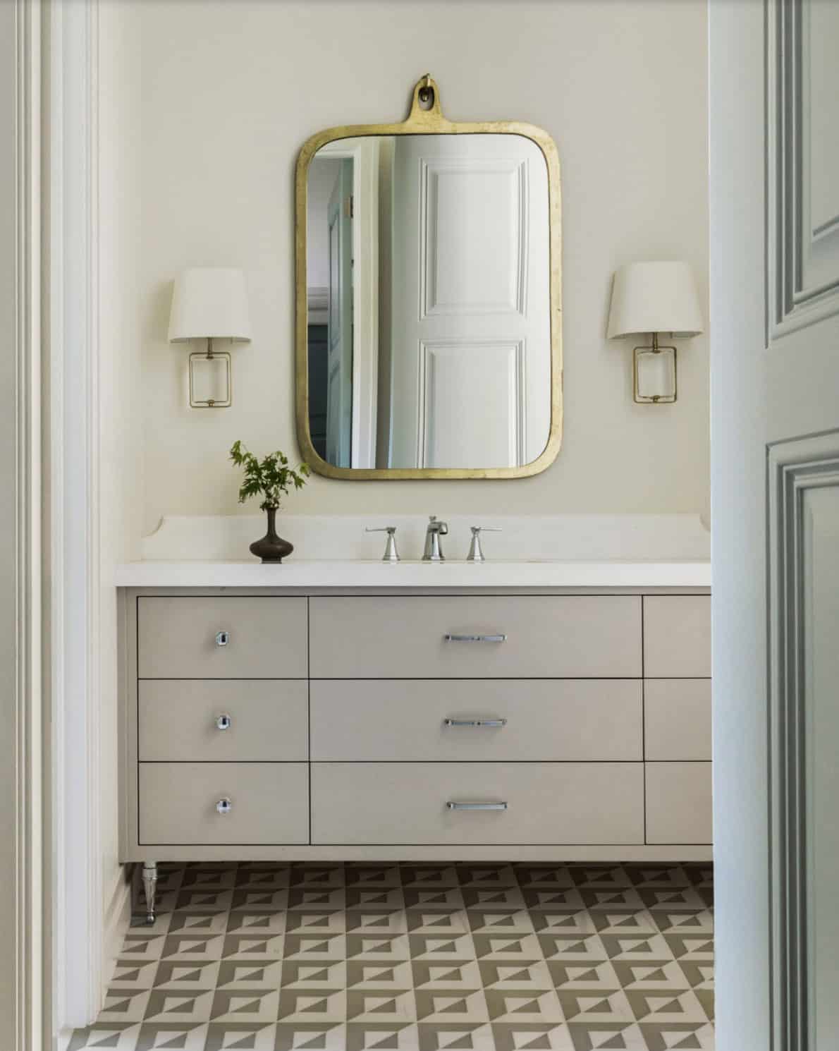 Guest bathroom vanity with taupe drawer cabinet, gold-framed mirror, brass sconces, and geometric tile floor