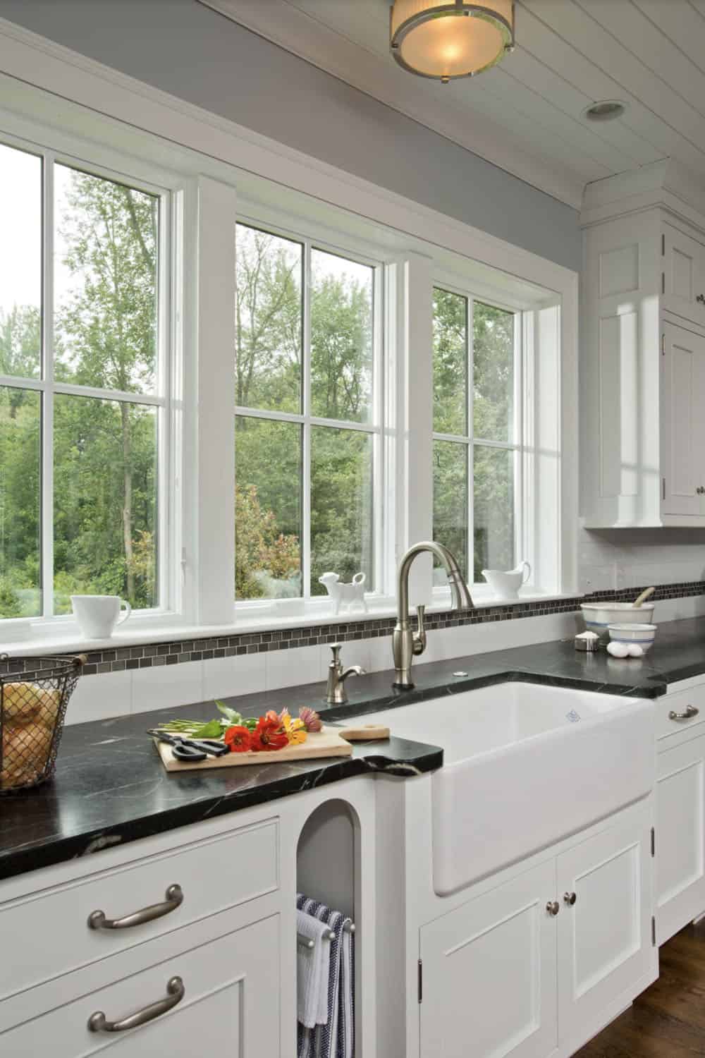 Farmhouse sink with black marble countertop and large windows overlooking wooded yard