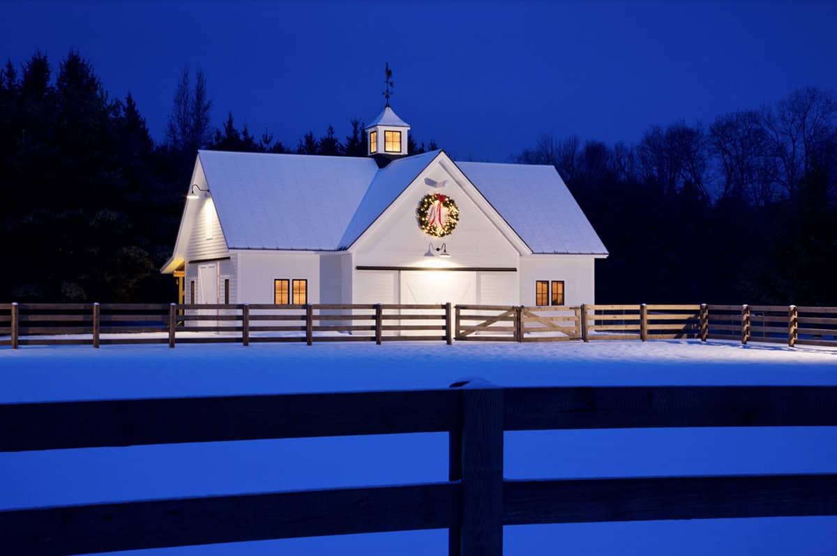 White barn with Christmas wreath and cupola at night in a snowy fenced paddock