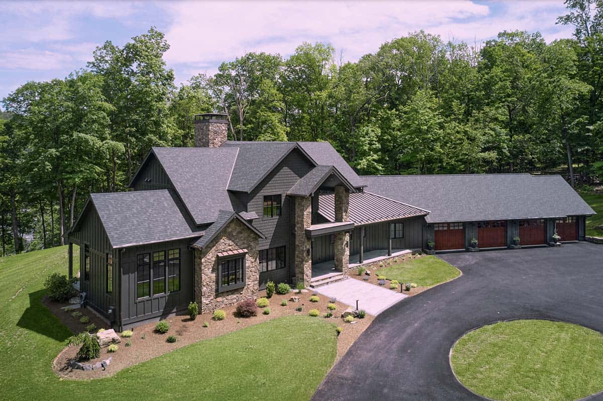 Aerial view of gray craftsman home with stone chimney, circular driveway, and forest backdrop