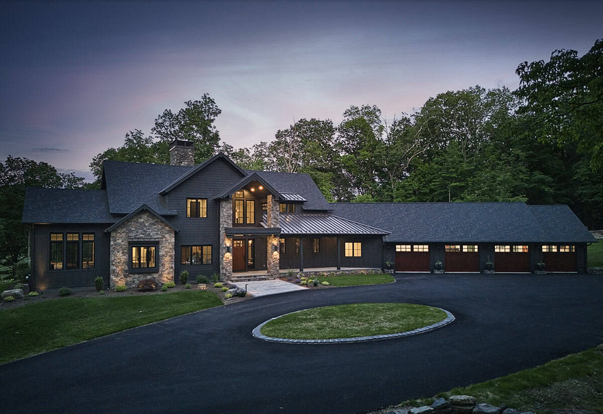 Dark craftsman home at dusk with glowing windows, circular driveway, and wood garage doors