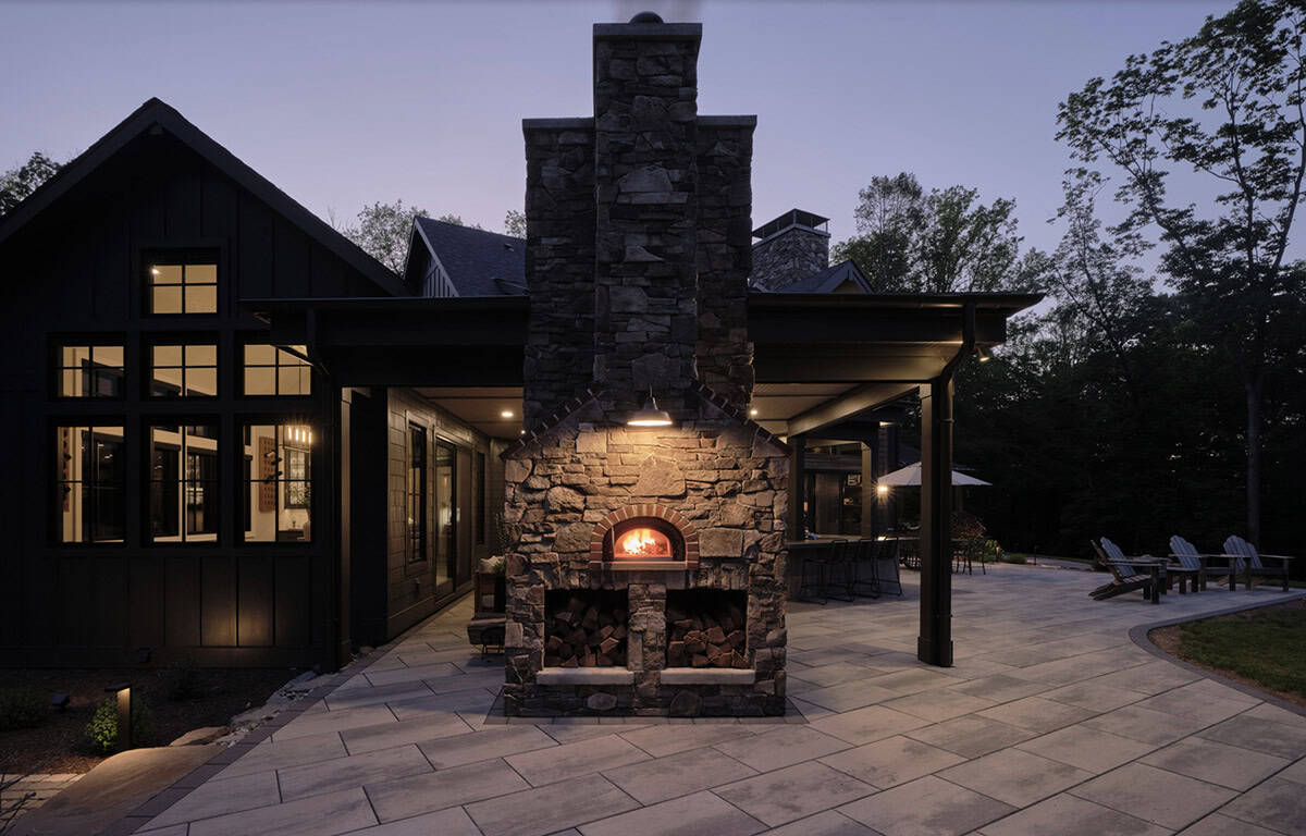 Stone outdoor pizza oven glowing at night on patio with covered bar area beyond