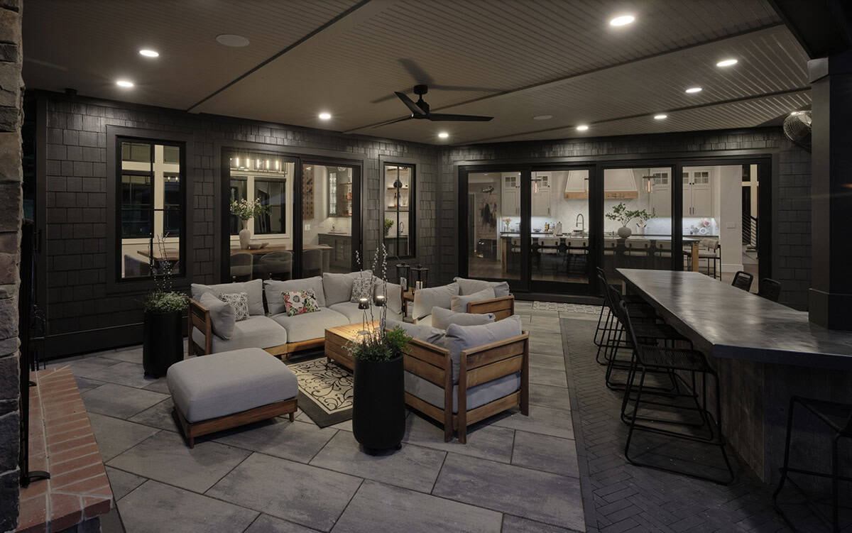 Covered outdoor living space at night with sectional seating, bar, and kitchen views through glass doors