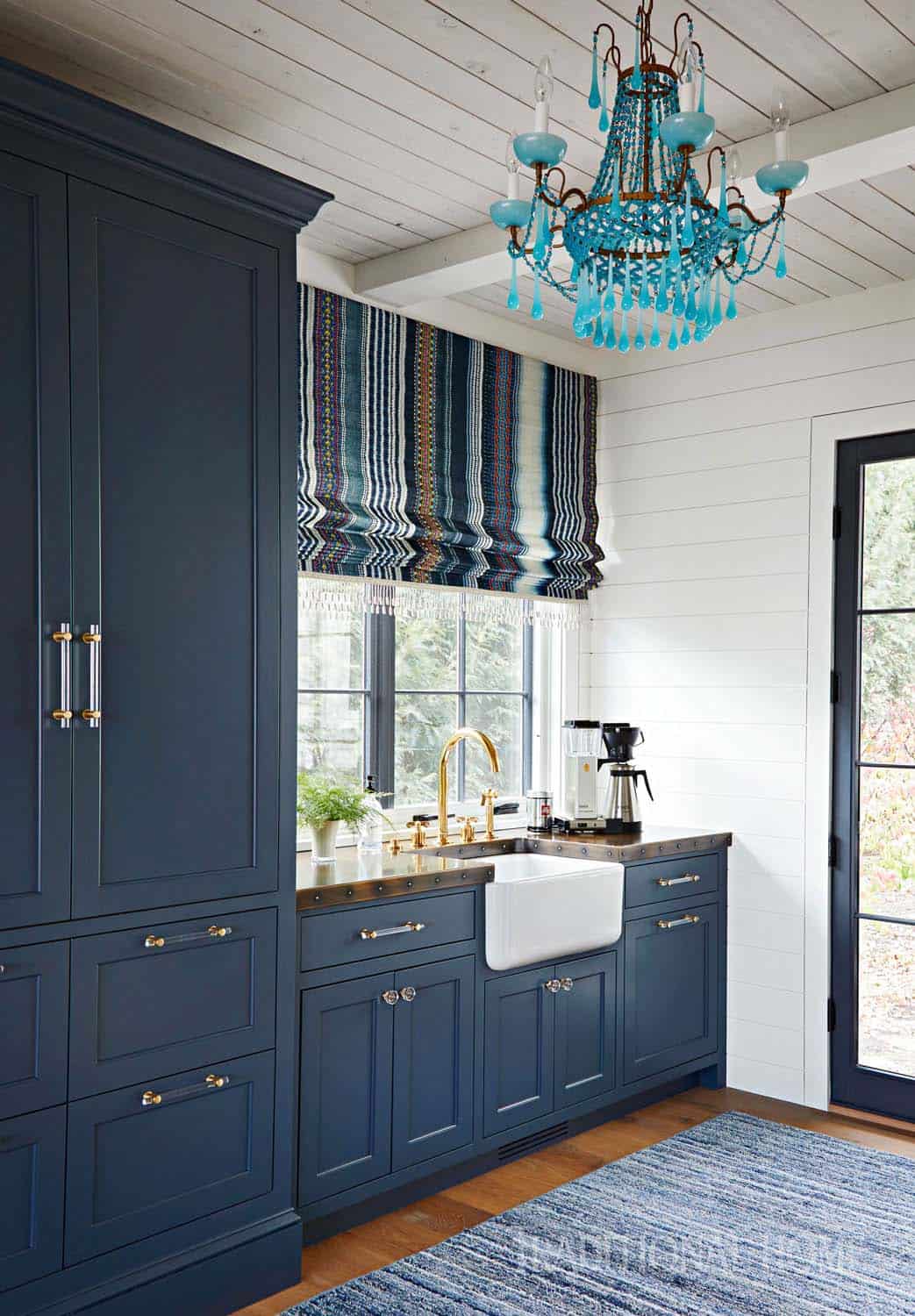 Navy blue butler's pantry with farmhouse sink, brass fixtures, striped roman shade, and turquoise beaded chandelier