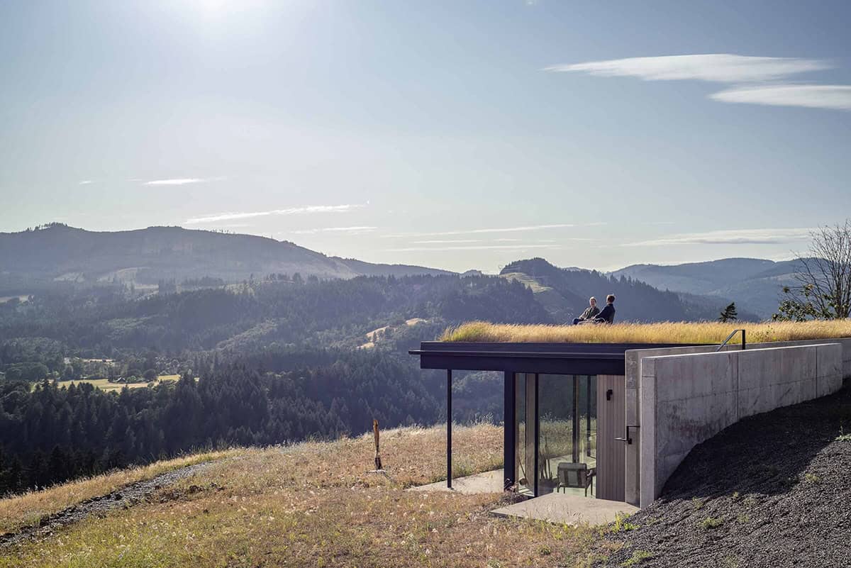 Concrete art studio bunker with grass roof and two people seated overlooking forested hills