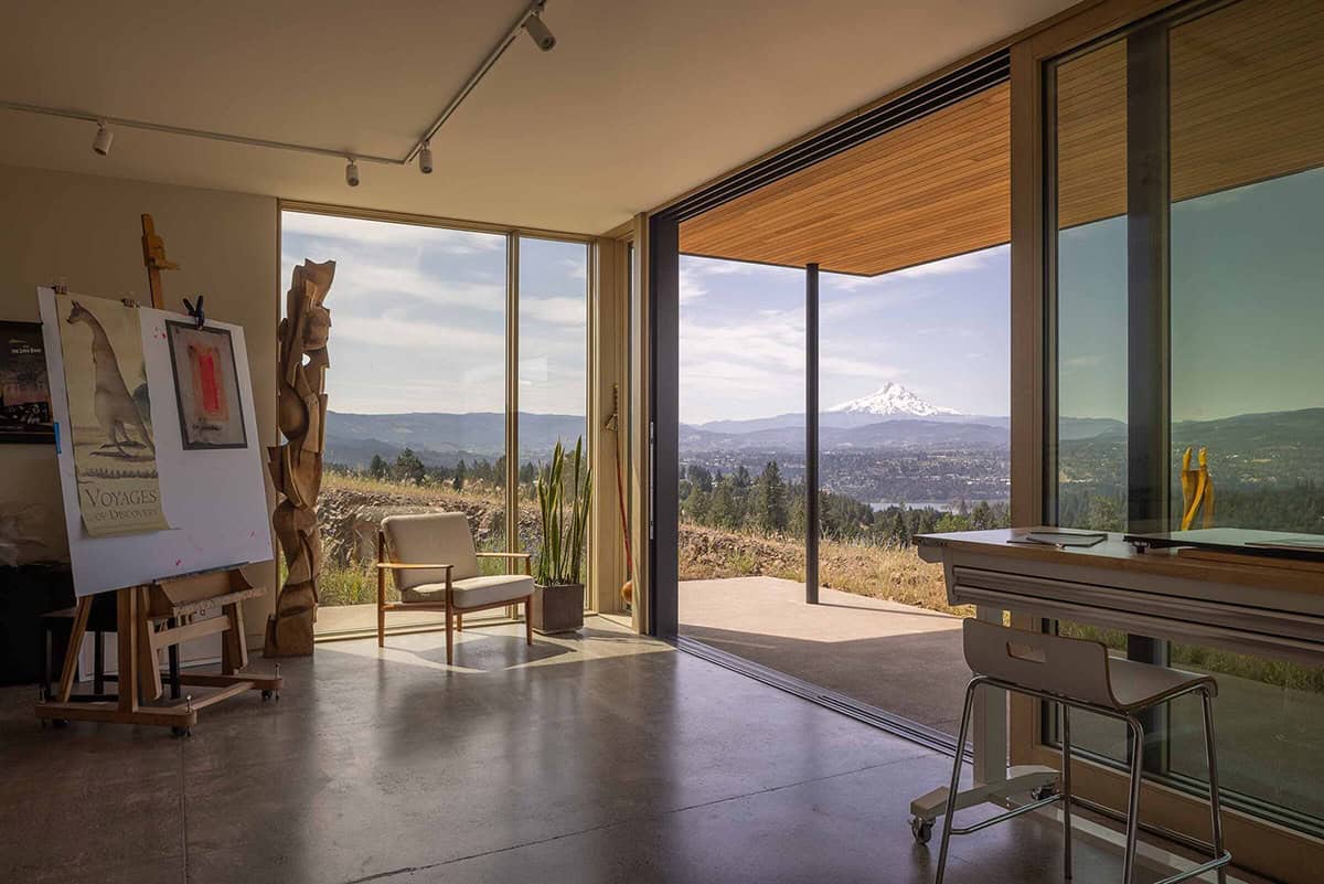 Artist's studio with polished concrete floors, easel, wood sculptures, and corner glass walls framing a mountain