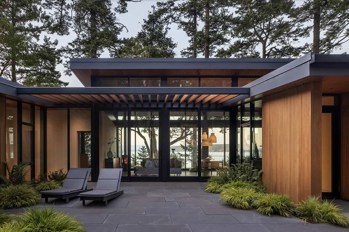 Courtyard view of modern Pacific Northwest home with floor-to-ceiling glass walls, cedar cladding, and slate lounge chairs
