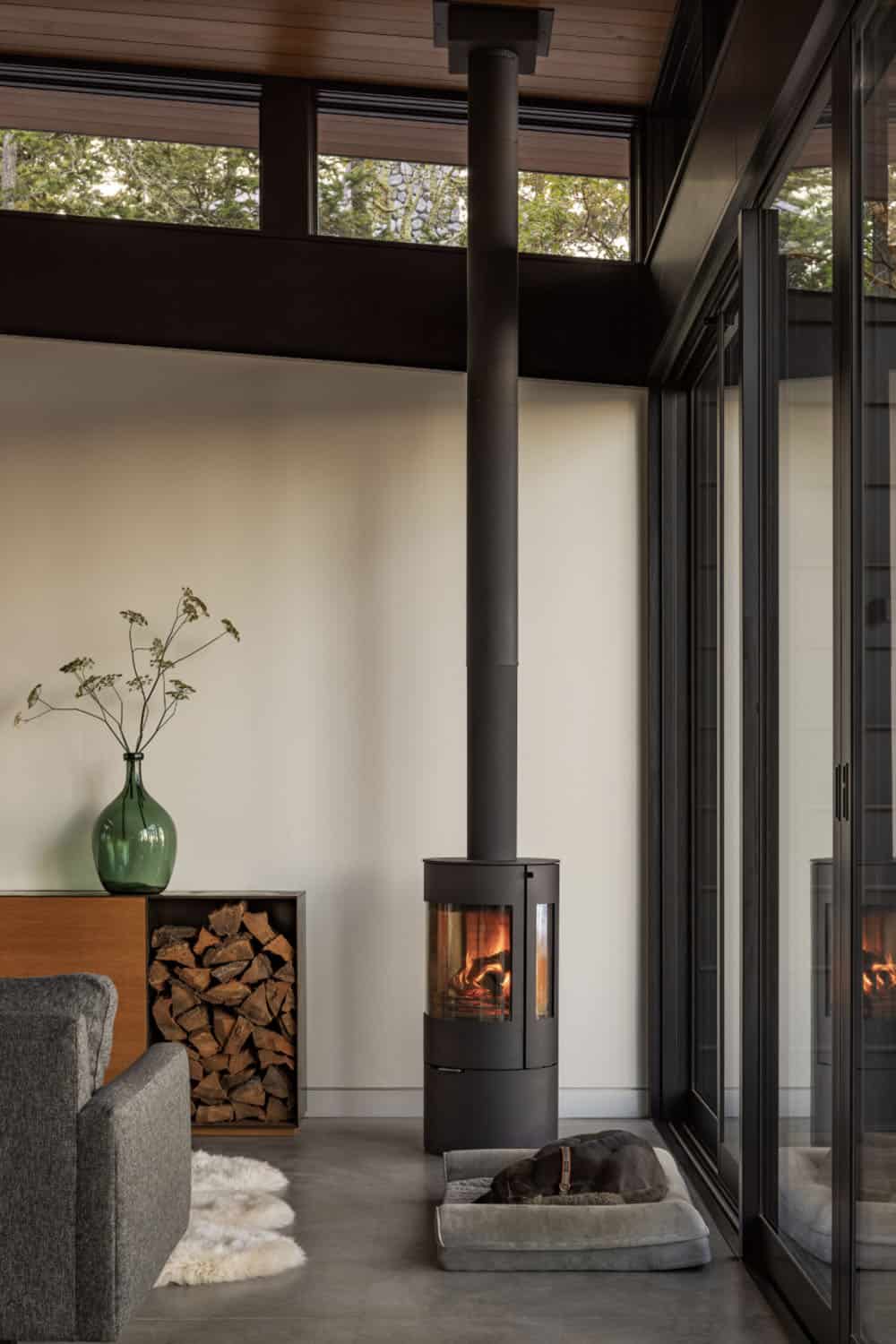 Cylindrical black freestanding wood stove with floor-to-ceiling pipe, beside built-in wood storage cabinet