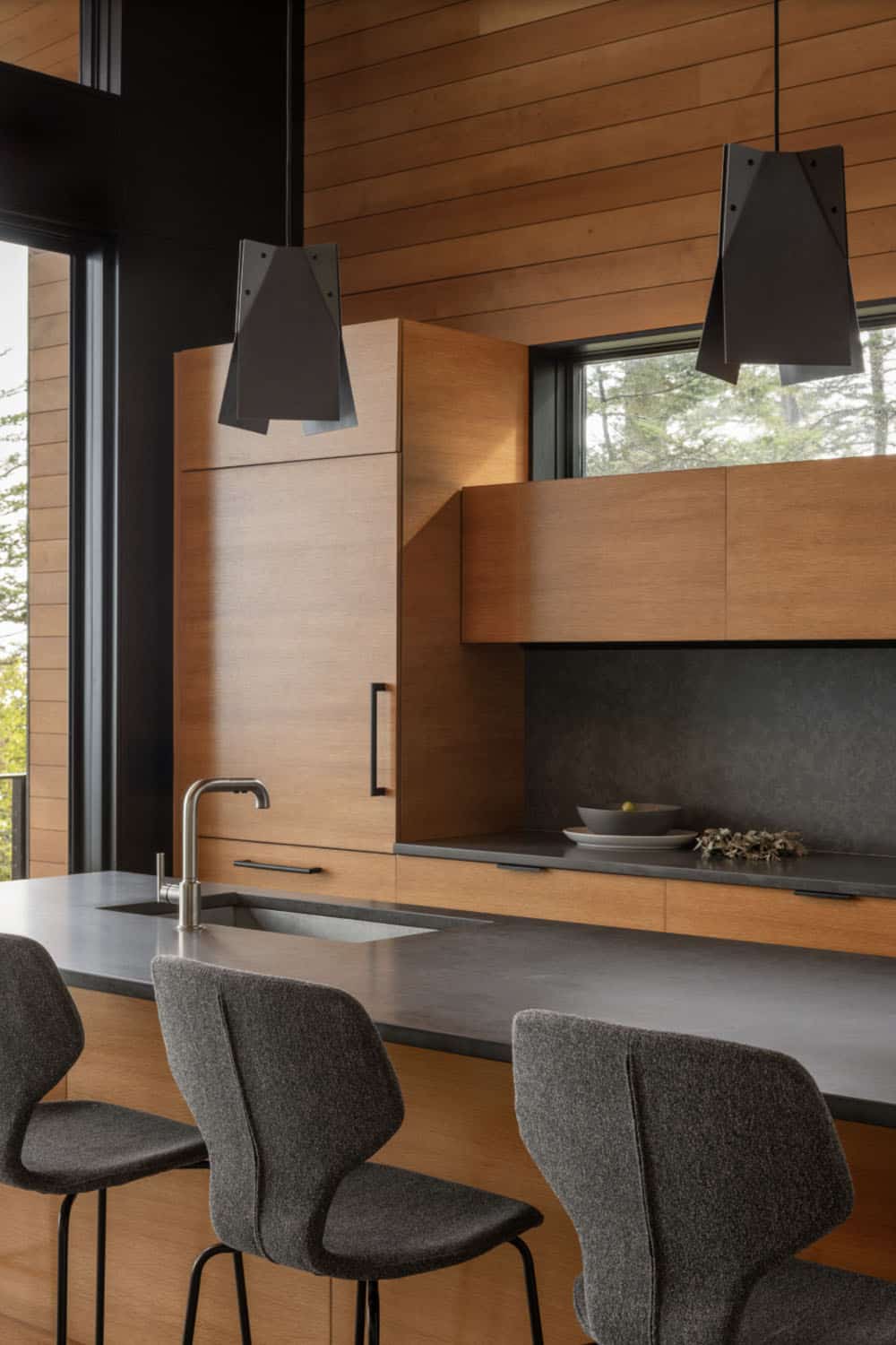 Kitchen island with dark stone countertop, gray boucle bar stools, and tall cedar cabinetry with angular pendants