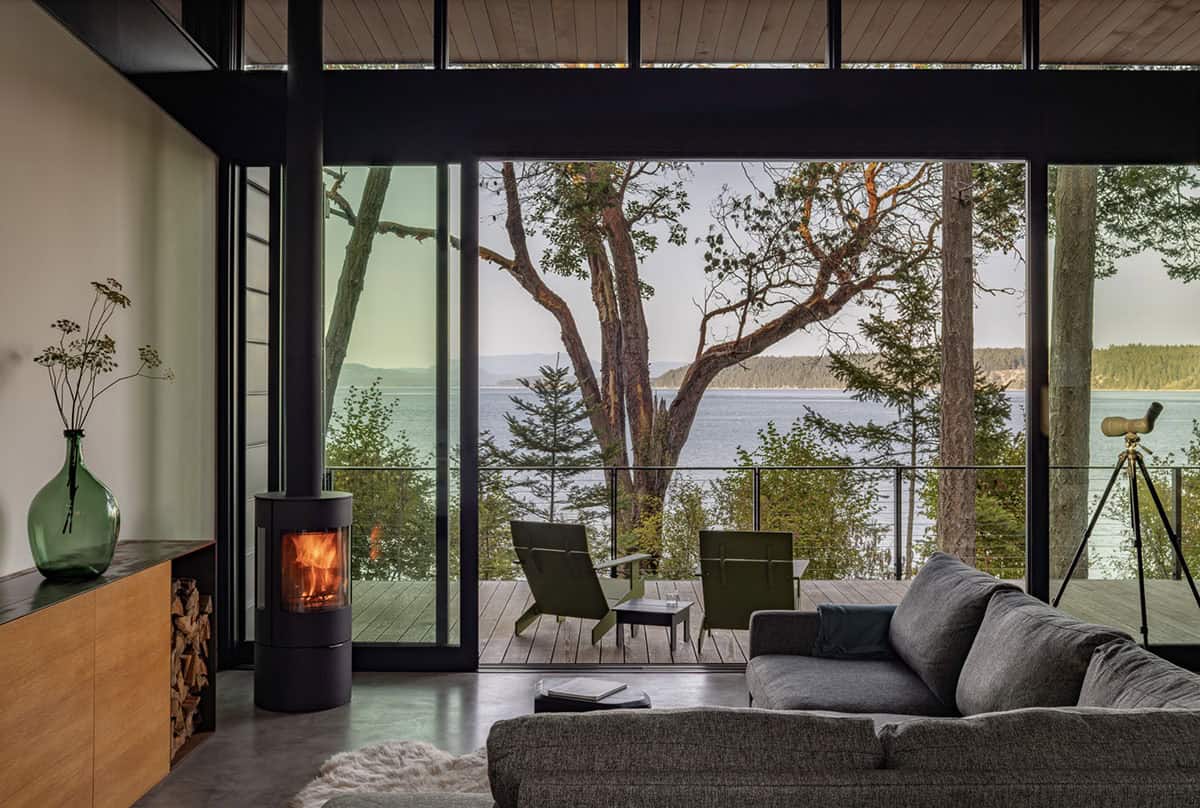 Living room with gray sectional, freestanding wood stove, and expansive glass wall opening to forested waterfront deck