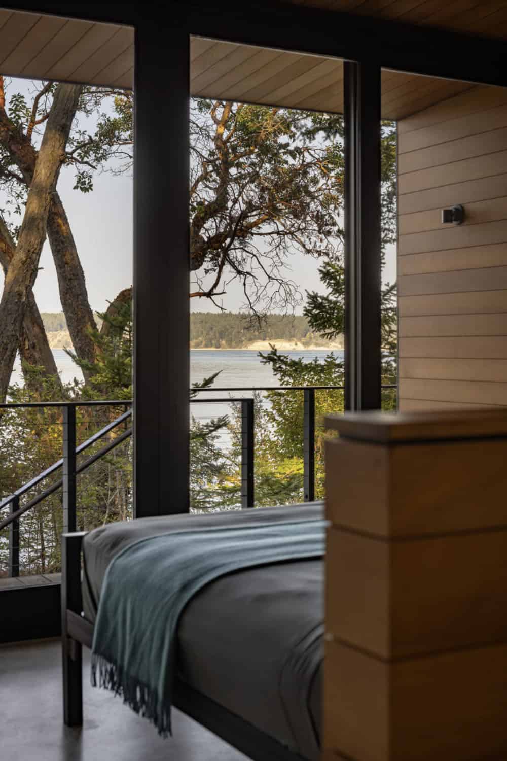 Bedroom corner with metal bed frame, teal throw blanket, and corner glass walls framing water and tree views