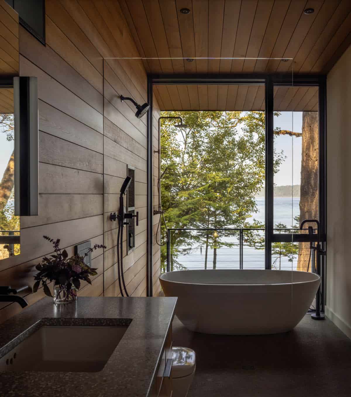 Primary bathroom with freestanding white soaking tub, black fixtures, wood plank walls, and floor-to-ceiling water view window
