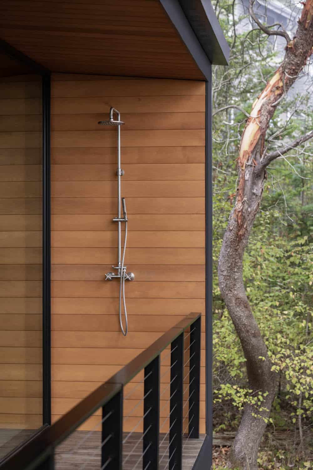 Outdoor shower with polished chrome fixtures mounted on cedar-clad exterior wall beside forested deck railing