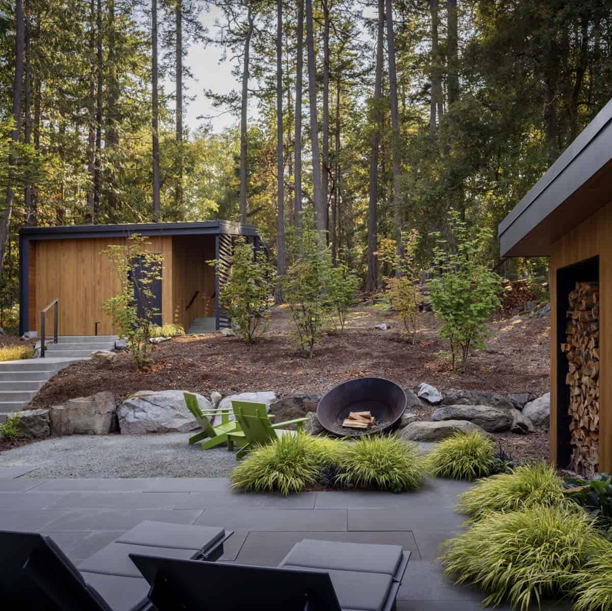 Landscaped outdoor area with fire pit bowl, green Adirondack chairs, ornamental grasses, and separate garage structure among pines