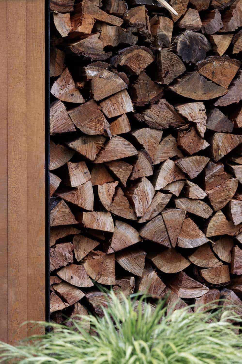 Close-up of split firewood stacked floor-to-ceiling in outdoor cedar storage cubby with ornamental grass in foreground
