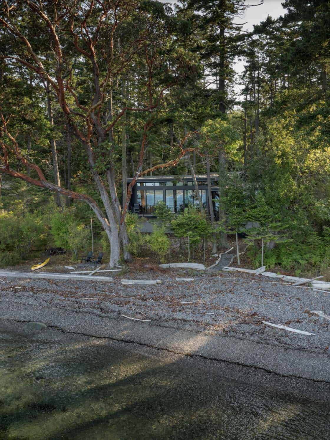 Waterfront view of minimalist black-framed glass residence set into forested hillside above rocky San Juan Island shoreline