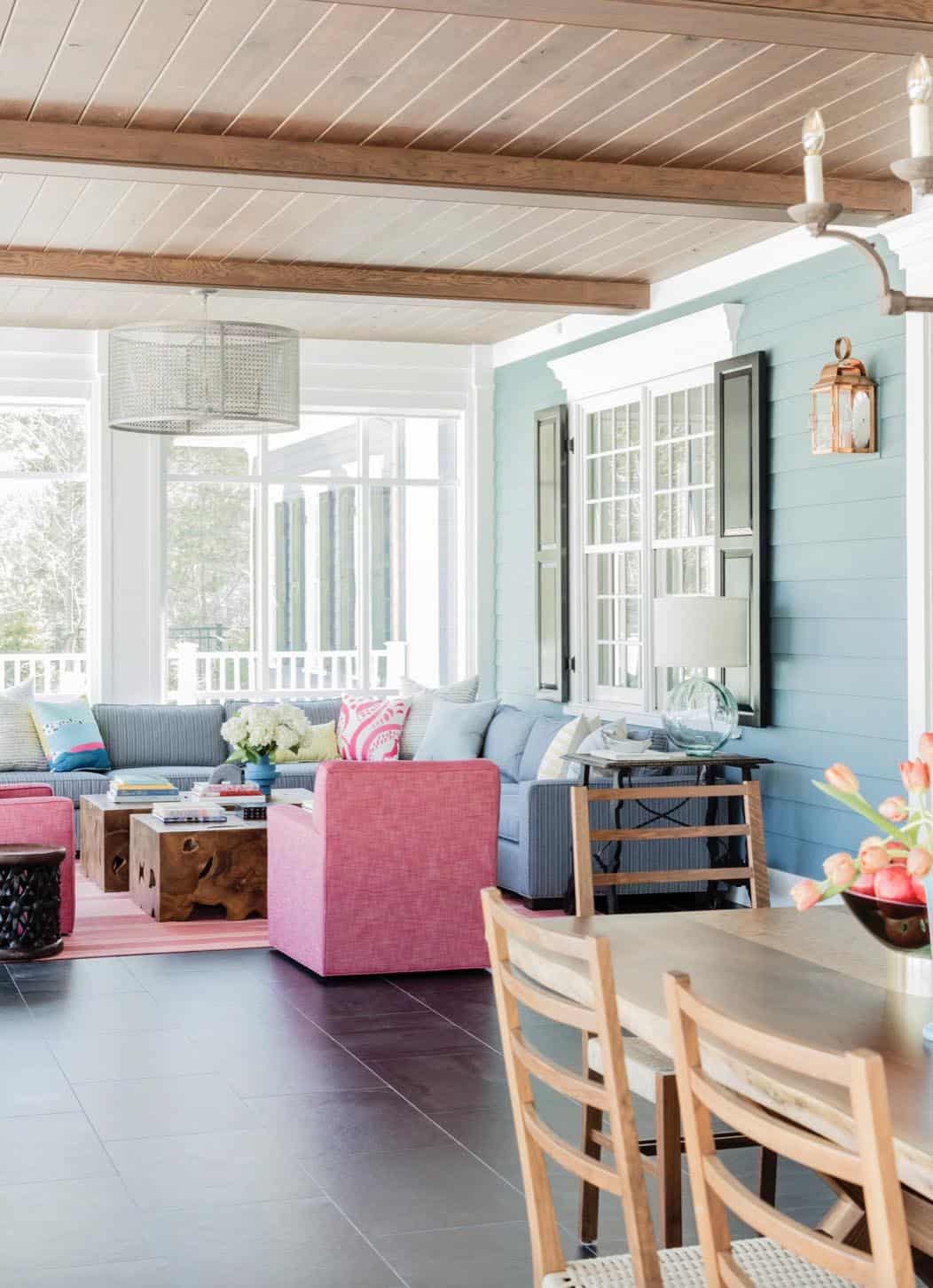Screened porch with blue striped sectional, pink armchair, teak coffee table, and blue shiplap