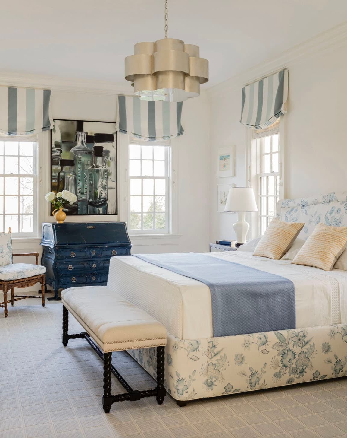 Primary bedroom with blue floral upholstered bed, striped roman shades, and sculptural chandelier