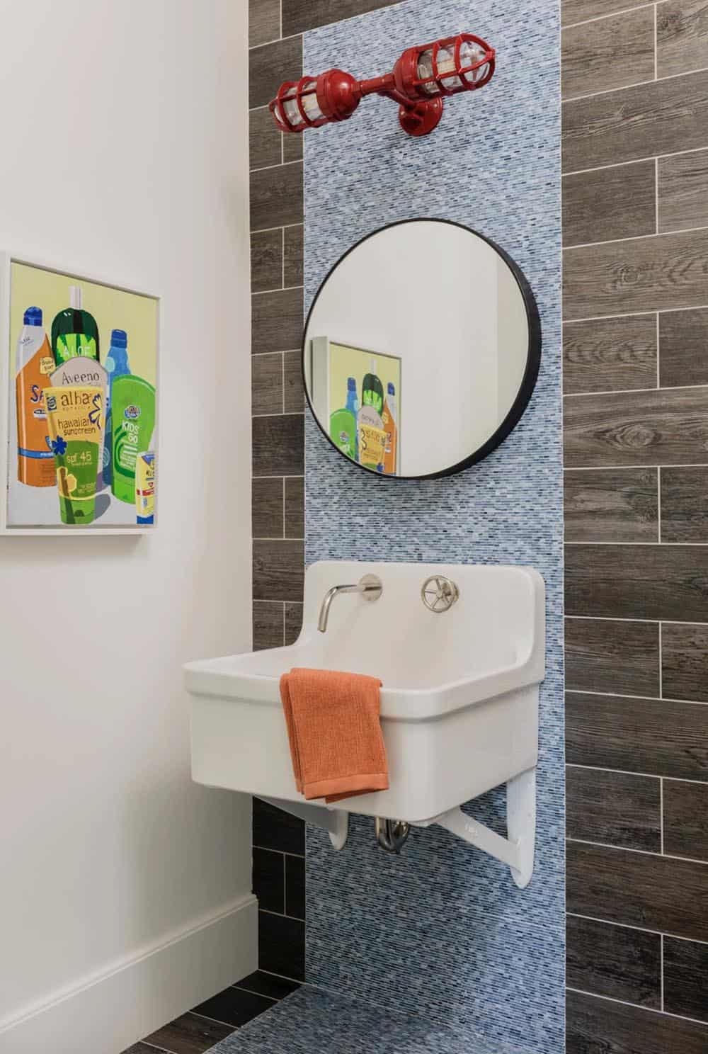 Kids' bathroom with blue mosaic tile accent, wall-mounted sink, round black mirror, and red cage sconce