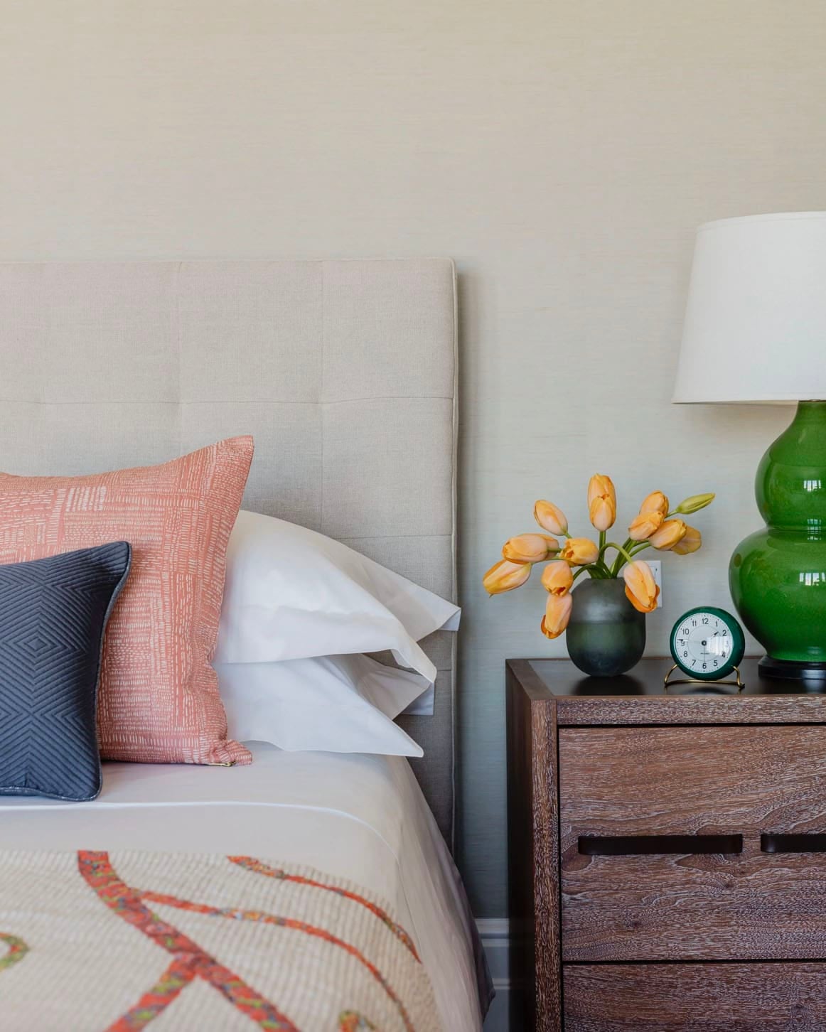 Bedroom nightstand detail with green ceramic lamp, yellow tulips, and coral throw pillow