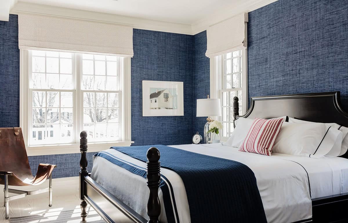 Navy grasscloth wallpaper bedroom with black four-poster bed, red striped pillow, and white roman shades