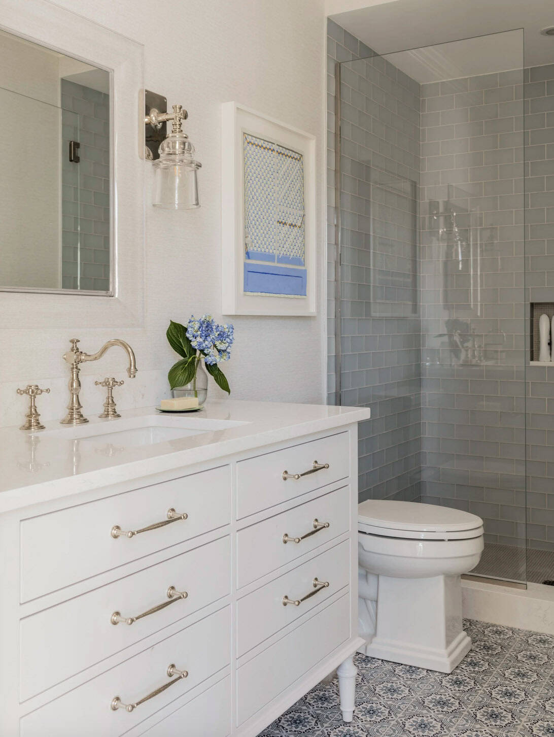 White bathroom with nickel fixtures, blue and white patterned floor tile, gray subway tile shower, and glass sconce
