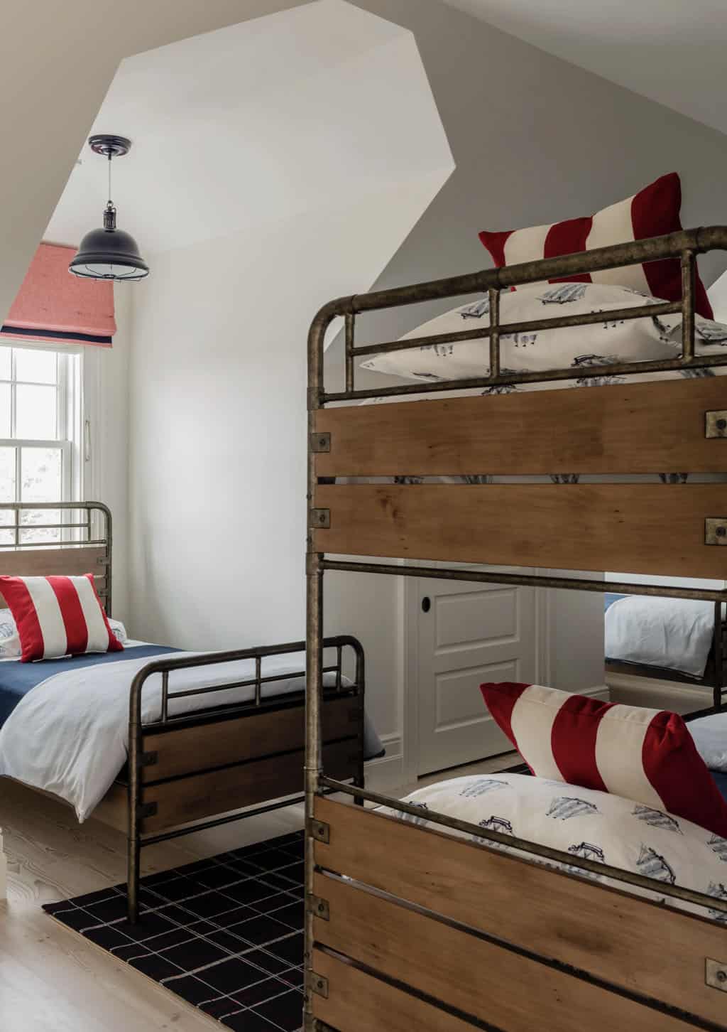 Attic bunk room detail with pipe-and-plank bunk beds, red and white striped pillows, and dormer window