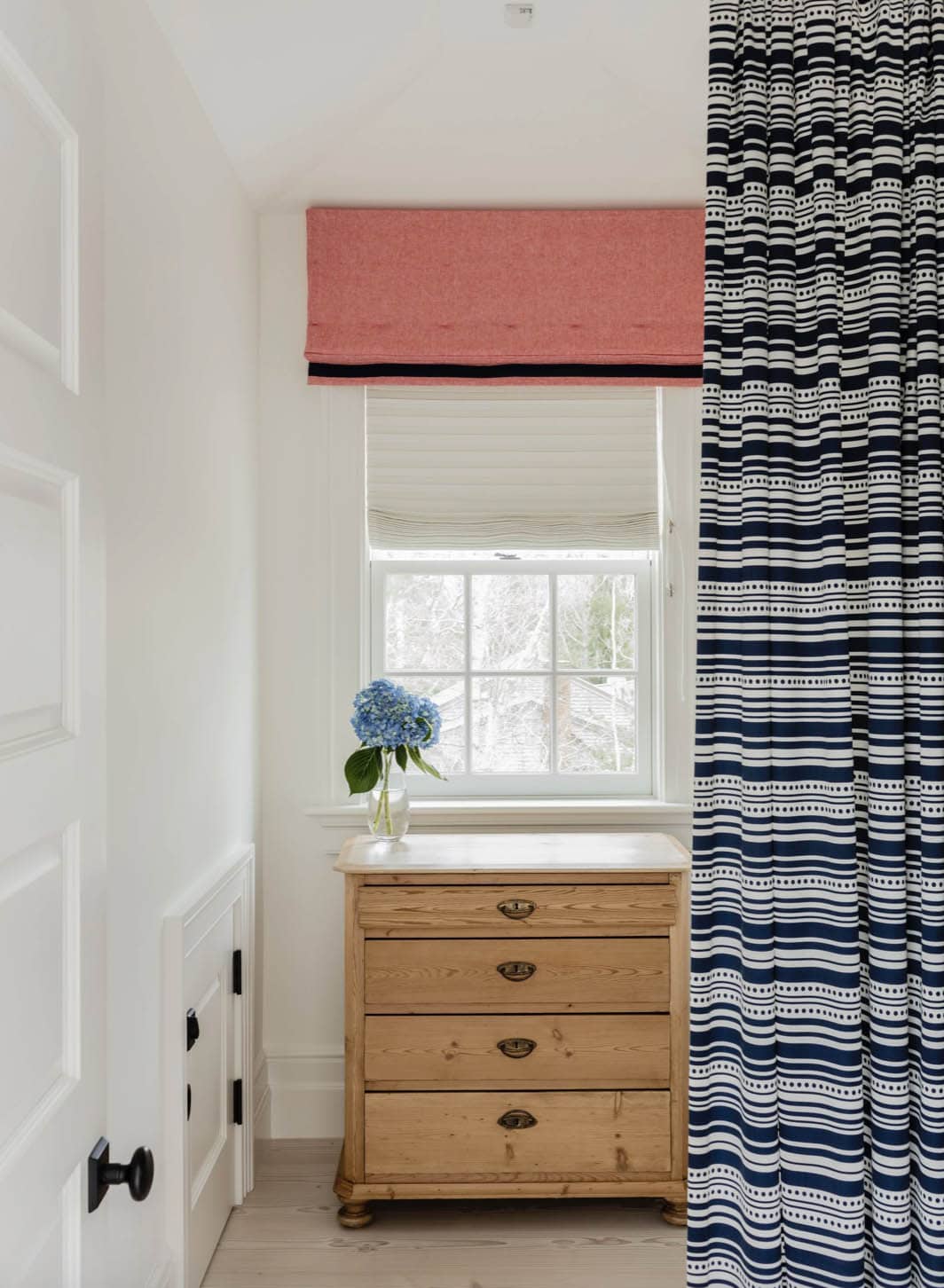 Bedroom corner with antique pine dresser, coral roman shade, and bold navy striped drape