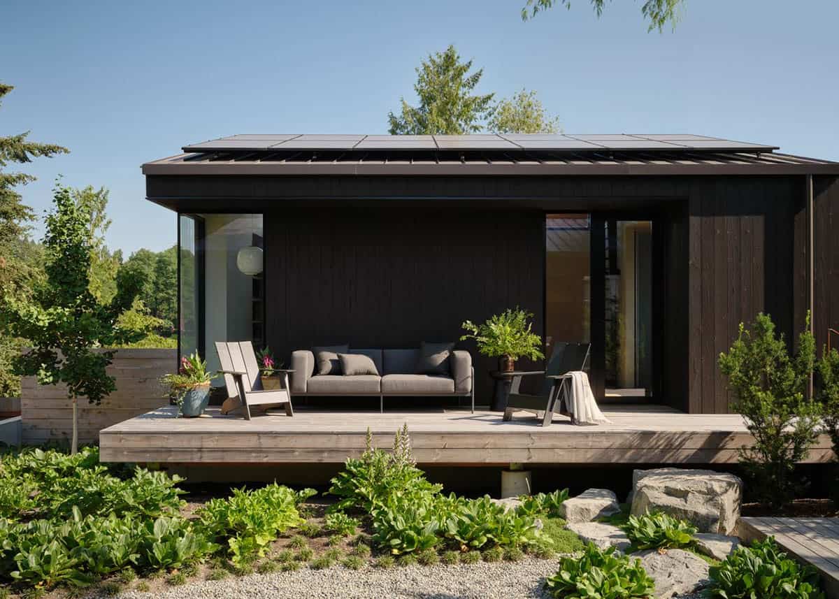 Dark-stained wood exterior with flat roof solar panels and covered deck with outdoor seating amid lush garden