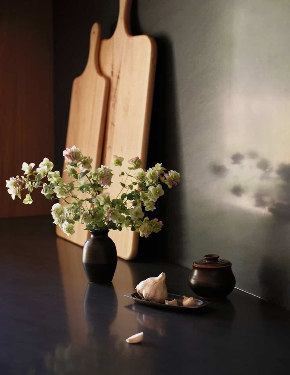 Dark kitchen counter vignette with small vase of blooms, cutting boards, and ceramic pot