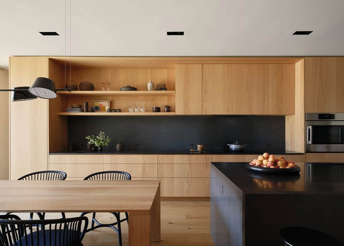 White oak kitchen cabinetry with black stone countertops, open shelving, and dining table