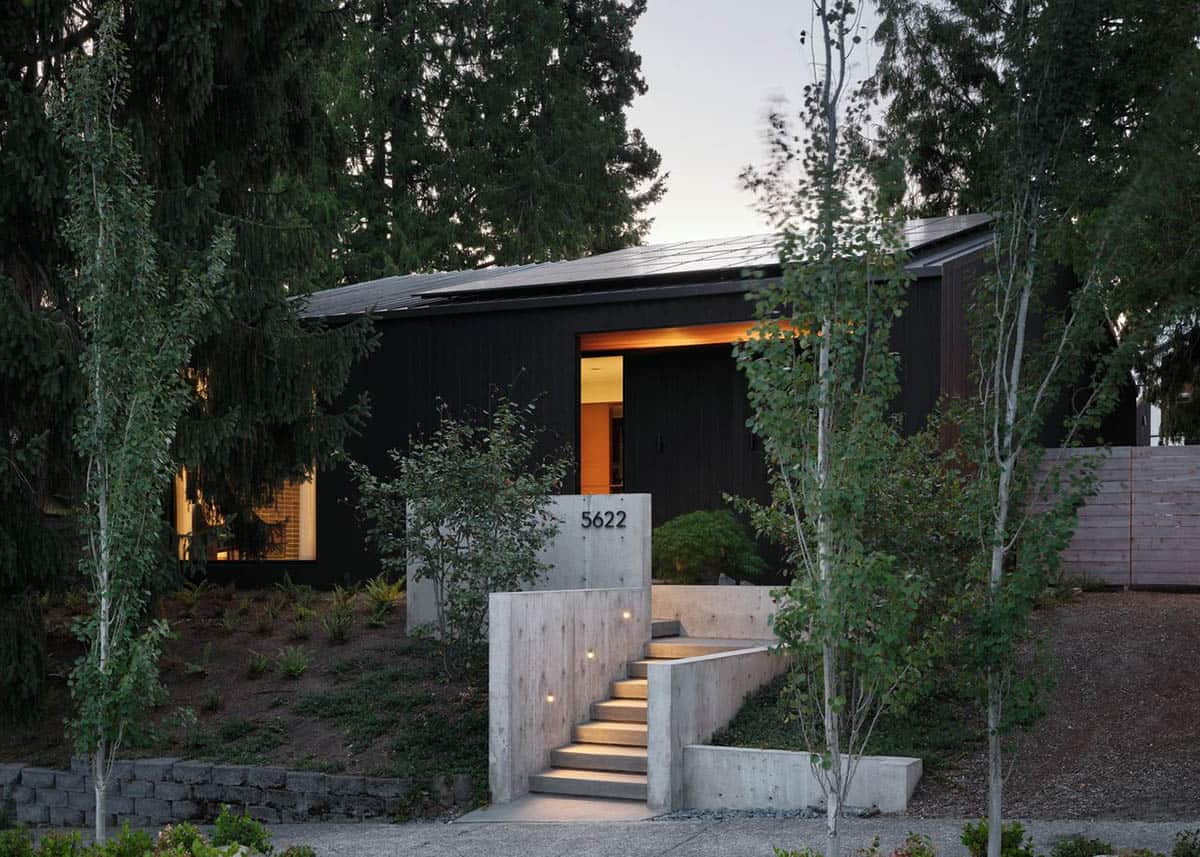 Dusk street view of dark wood house with lit concrete entry stairs, solar roof, and surrounding trees