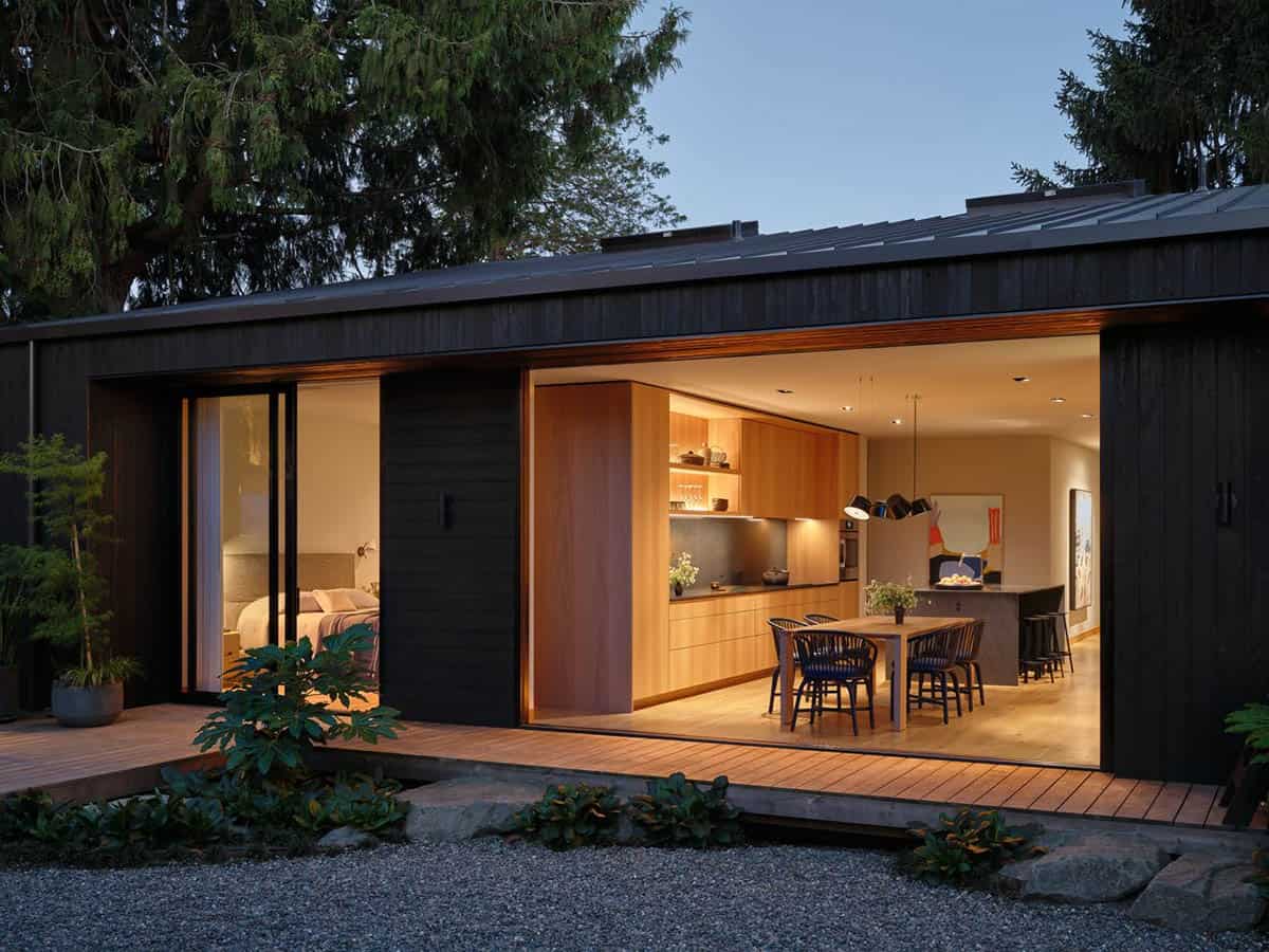 Twilight exterior of dark-clad home with wide open sliding doors revealing warmly lit kitchen and dining interior