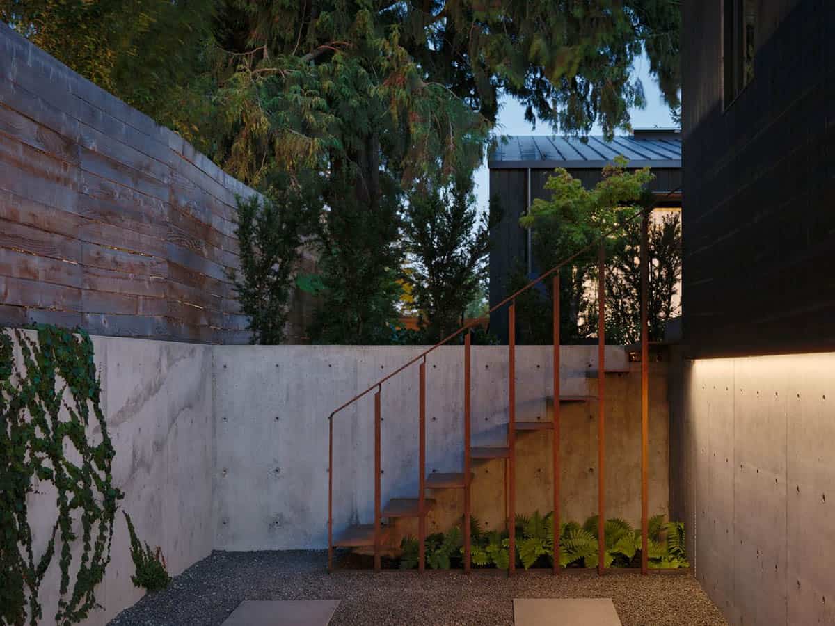 Dusk view of concrete retaining walls with Corten steel stair railing, ferns, and ivy beside dark wood exterior